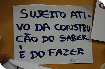 Galpão rústico com pessoas sentadas no chão em roda e uma pessoa em pé falando. 
