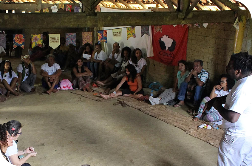 Galpão rústico com pessoas sentadas no chão em roda e uma pessoa em pé falando. 