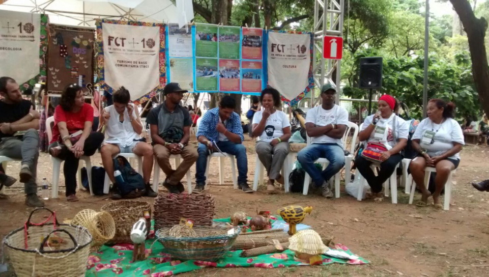 Foto com várias pessoas ligadas ao fórum de comunidades tradicionais estão sentadas, lado a lado, tendo ao fundo estandartes com fotos e cartazes pendurados, como um mural, e à frente objetos tradicionais produzidos pela comunidade, como cestos de vime. 
