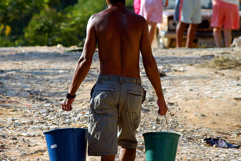 Homem sem camisa, no sol, carregando dois baldes com água. 